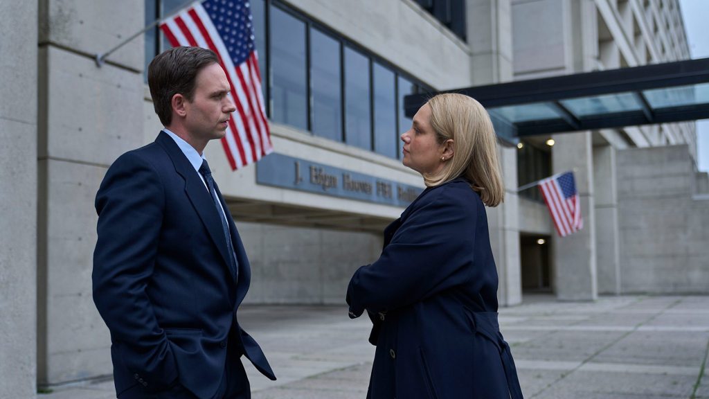 A woman stands in front of man with arms folded.