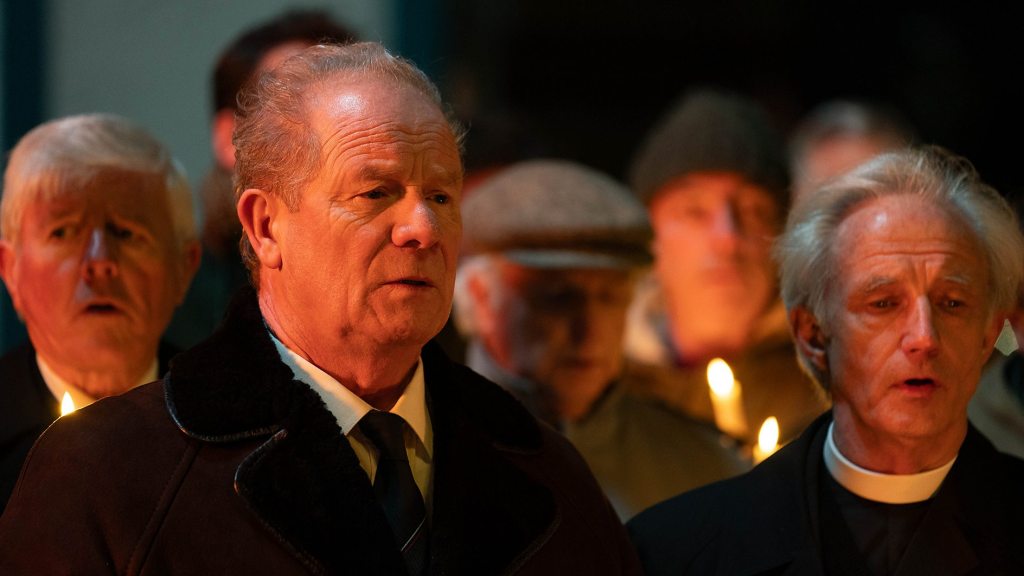 A man stands next to another man at a funeral.